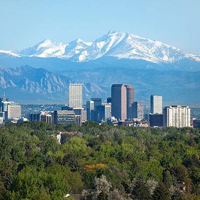 Snow covered Longs Peak, part of the Rocky Mountains stands tall in the background with green trees and the Downtown Denver skyscrapers as well as hotels, office buildings and apartment buildings filling the skyline.